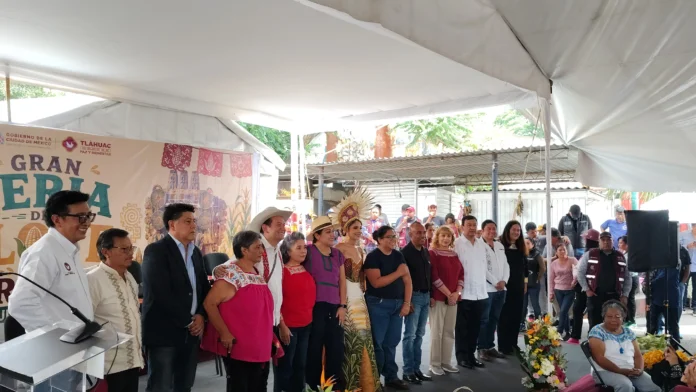 Group of people standing together on a stage under a white tent at a cultural festival; several wear traditional clothing and hats, with a decorative banner in the background.