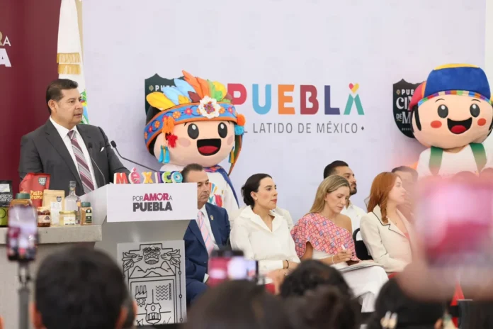 Puebla será sede del Tianguis Turístico México 2027 Man in a suit speaks at a podium during a Puebla event, with colorful mascots and a panel of people seated behind him.